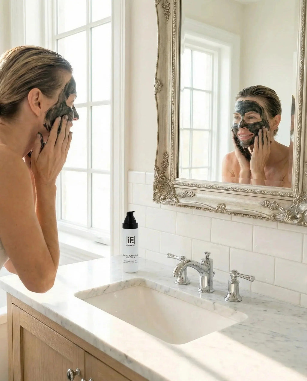 Woman applying detox black cleanser face mask in bright bathroom with mirror and skincare bottle
