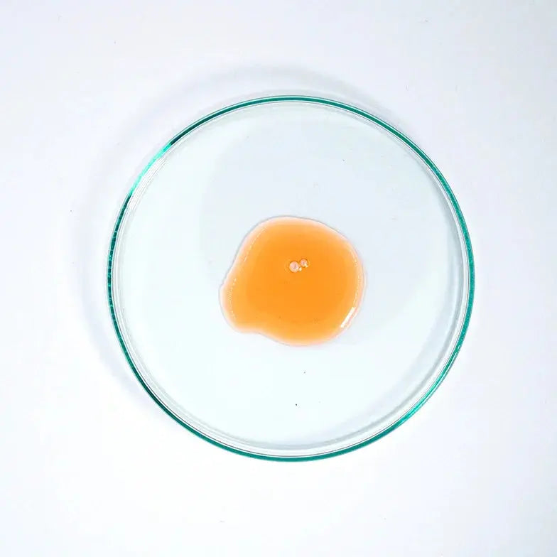 Golden glycolic serum swatch in clear petri dish on white background, skincare texture