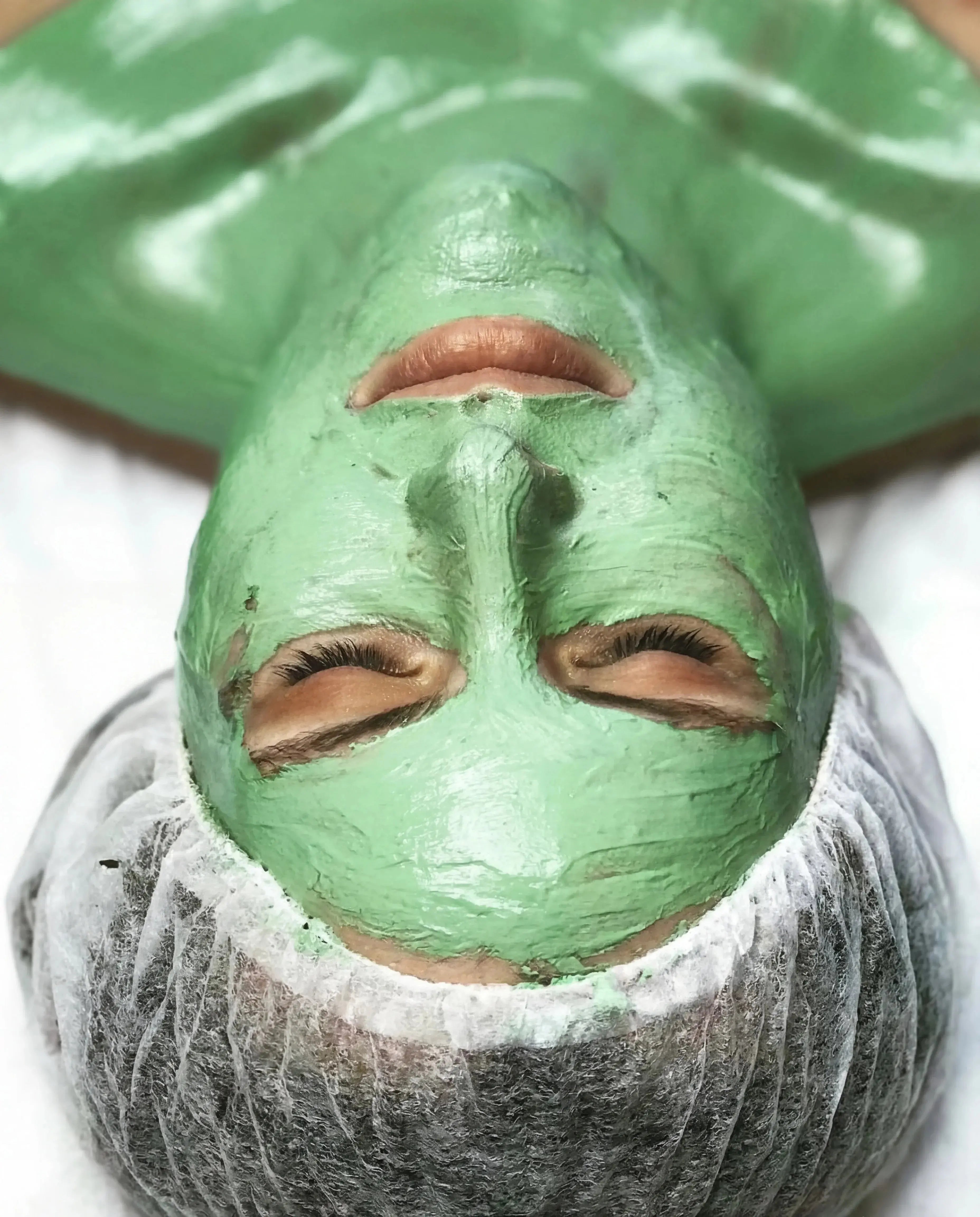 Woman with closed eyes wearing a green clay facial mask and a hair cap, spa skincare treatment
