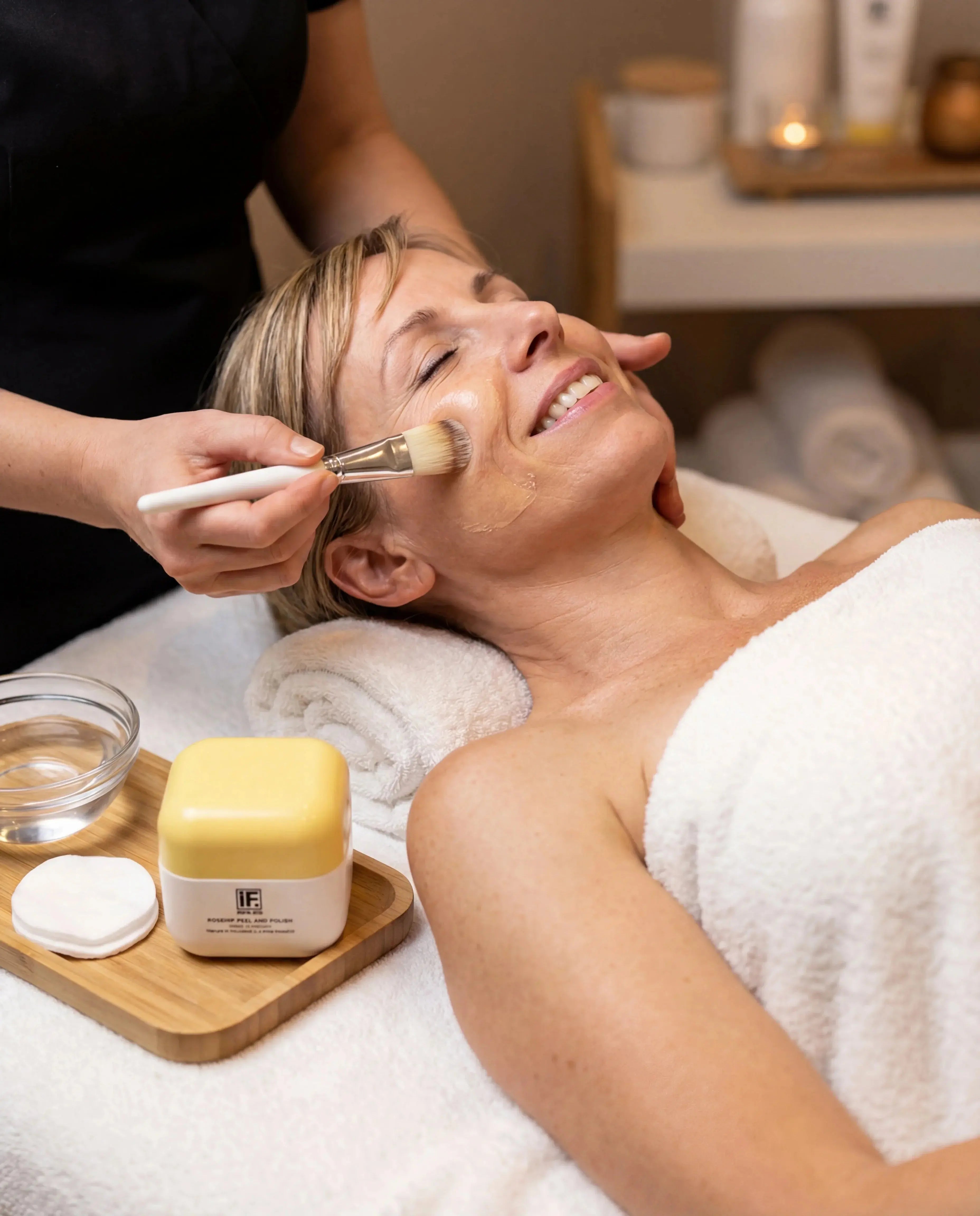 Woman enjoying rosehip peel and polish facial treatment at a spa with skincare products visible.