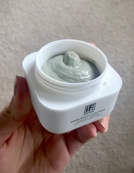 Hand holding open jar of green clay face mask, creamy texture visible inside white container